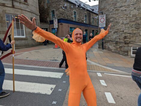 A person in an orange full-body suit stands with arms outstretched on a street near stone buildings, with people and a "no entry" sign in the background.