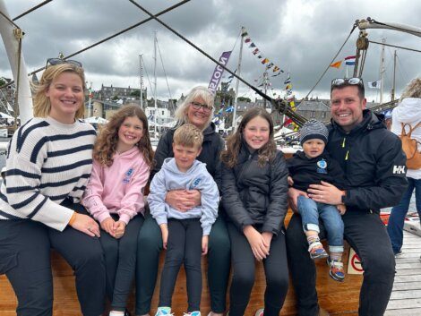 A group of six people, four children and two adults, sit on a boat in a marina. It is a cloudy day and sailboats are visible in the background.