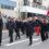 A group of people in navy uniforms march in a parade, some waving, with a Norwegian flag visible and a sign reading "Statsraad Lehmkuhl Norway." Spectators stand behind a low wall in the background.