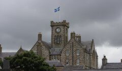 A historic stone building with a clock tower and a flag on top, set against a cloudy sky.