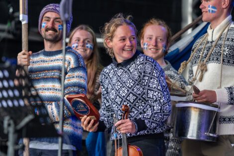 A group of people wearing patterned sweaters and face paint hold musical instruments, including a violin and a drum, smiling and standing together at an event.