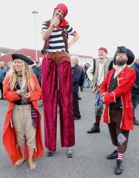 A group of people in pirate costumes, including one person on stilts, are standing together outdoors among a crowd.