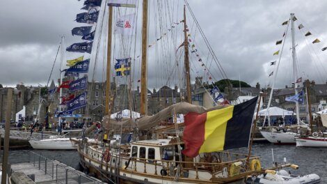 A wooden sailboat with a Belgian flag docked at a marina, surrounded by other boats and flags, under a cloudy sky.