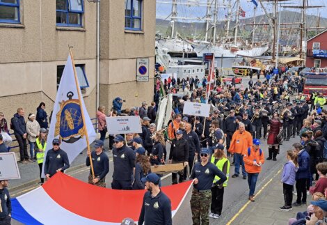 A parade with participants in navy uniforms, some carrying flags and signs. Spectators are lined up on both sides of the street. Sailboats are visible in the background.