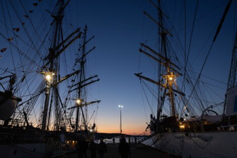 A twilight scene with silhouettes of people walking on a dock beside two tall sailing ships with illuminated masts against a fading sunset.