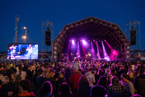 A crowd gathers in front of a large outdoor stage with purple lighting under a dark sky. A live band performs, and a screen to the left displays the action on stage.