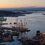 Aerial view of a harbor at sunset with several docked ships, including a tall-masted sailboat, surrounded by buildings and calm water.