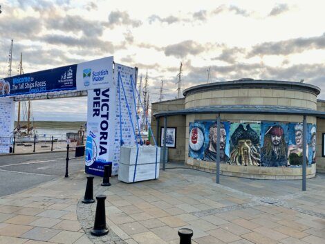 Entrance arch to Victoria Pier for The Tall Ships Races, with building decorated with pirate-themed murals. Overcast sky and ships visible in the background.