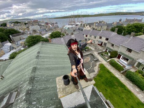 Person dressed as a pirate taking a selfie atop a roof with a town and waterfront in the background on a cloudy day.