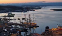 Aerial view of a harbor at sunset with several docked ships, including a tall-masted sailboat, surrounded by buildings and calm water.