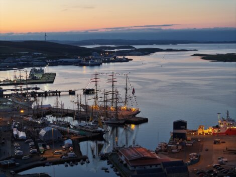 Aerial view of a harbor at sunset with several docked ships, including a tall-masted sailboat, surrounded by buildings and calm water.