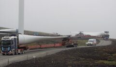 Heavy trucks transport large wind turbine blades on a gravel road in a foggy landscape. A service van is parked nearby.