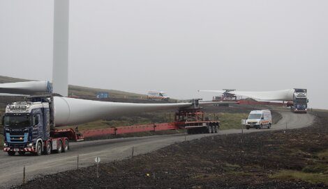 Heavy trucks transport large wind turbine blades on a gravel road in a foggy landscape. A service van is parked nearby.