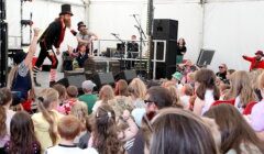 A performer dressed in a top hat and striped socks entertains a crowd of children and adults in a tented venue. Other performers and equipment are visible on stage.