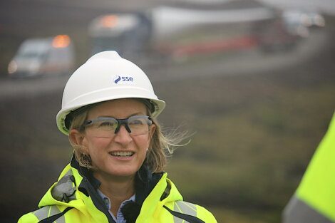 A person in a high-visibility jacket and hard hat with an "SSE" logo is outdoors, smiling. Several vehicles can be seen in the blurred background on a road.