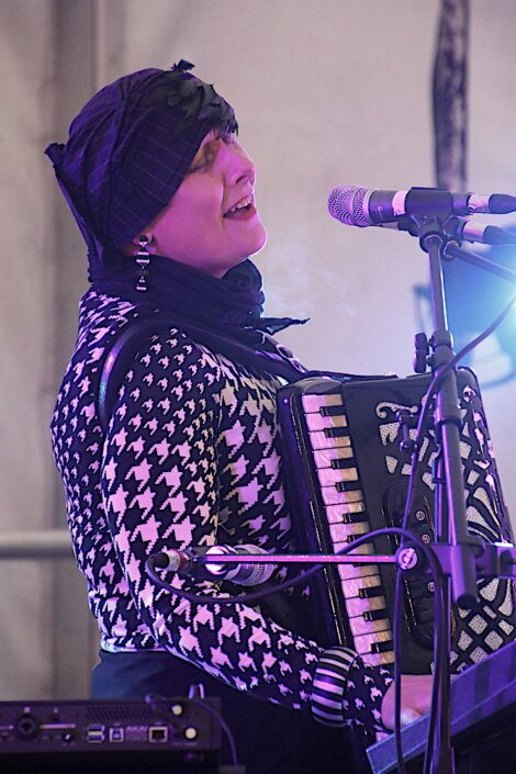 A person wearing a black and white patterned sweater and headscarf plays an accordion and sings into a microphone on stage.