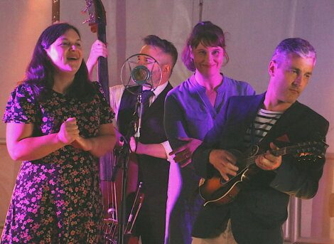 Four musicians are gathered, three dressed in vintage-style clothing and one playing a mandolin. A woman with a floral dress is singing, and a man plays an upright bass.