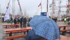 A person sits at a wooden bench holding a blue plaid umbrella at a maritime event with tall ships in the background, on a cloudy and rainy day. People are walking around, some also carrying umbrellas.