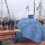 A person sits at a wooden bench holding a blue plaid umbrella at a maritime event with tall ships in the background, on a cloudy and rainy day. People are walking around, some also carrying umbrellas.