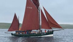 A green and black traditional sailing boat with brown sails, labeled LK 243, floats on calm waters with a few people onboard and land in the distant background.