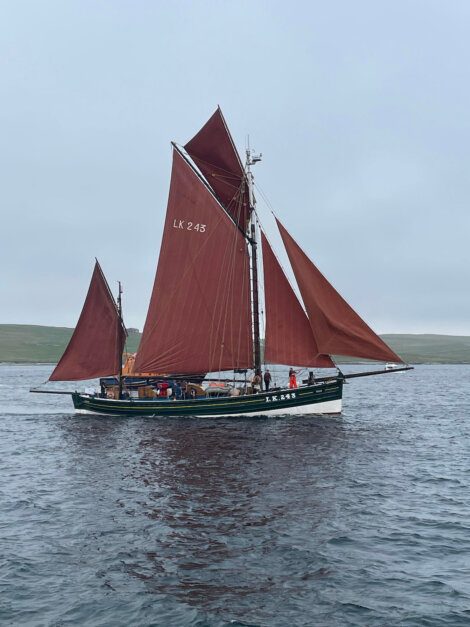 A green and black traditional sailing boat with brown sails, labeled LK 243, floats on calm waters with a few people onboard and land in the distant background.