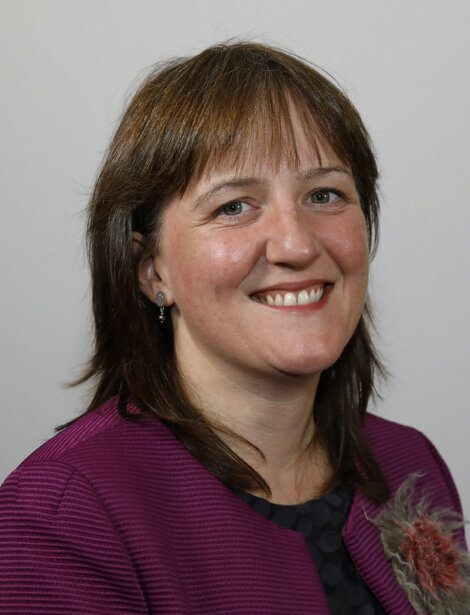 A person with straight brown hair, wearing a purple blazer and smiling, is posed against a plain grey background.