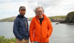 Two men stand outdoors on a rocky coastal landscape. One wears a blue jacket and cap, the other an orange puffy jacket. Both smile at the camera under a cloudy sky.