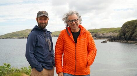 Two men stand outdoors on a rocky coastal landscape. One wears a blue jacket and cap, the other an orange puffy jacket. Both smile at the camera under a cloudy sky.