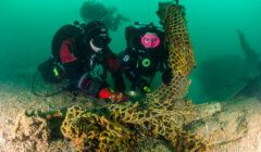 Two scuba divers in full gear remove a large fishing net from the ocean floor, with a buoy attached to the net for lifting.