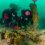 Two scuba divers in full gear remove a large fishing net from the ocean floor, with a buoy attached to the net for lifting.