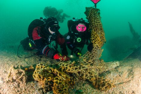 Two scuba divers in full gear remove a large fishing net from the ocean floor, with a buoy attached to the net for lifting.