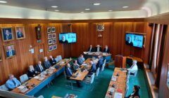 A group of people in formal attire sit around a U-shaped table in a wood-paneled room, conducting a meeting with some participants joining via video conference displayed on screens.