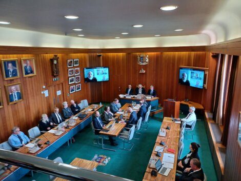 A group of people in formal attire sit around a U-shaped table in a wood-paneled room, conducting a meeting with some participants joining via video conference displayed on screens.