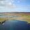 Aerial view of a coastal landscape with cliffs, green fields, scattered buildings, and a body of water under a blue sky with clouds.