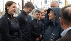 A woman in a raincoat is talking to a group of people in matching jackets. They appear to be outdoors near boats with various flags displayed.