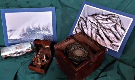 A display featuring two photographs, a ship in a bottle, a marine instrument in a wooden box, and a compass in a wooden case, set on a green fabric backdrop.