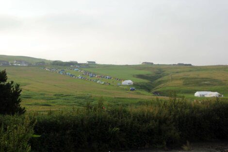 A grassy landscape with numerous colorful tents scattered across rolling hills. Several small buildings can be seen in the background. The sky is overcast.