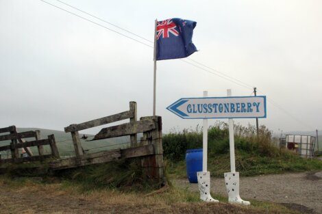 A flag waves on a post next to wooden fence. A sign labeled "Glusstonberry" with arrows points forward with two white rain boots placed by its base. Grass and a hazy sky can be seen in the background.