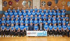 Group photo of Team Shetland in blue shirts, standing and seated in rows, holding a banner with sponsors "Malakoff Limited" and "Team Shetland." There are six plaques displayed on the wall behind them.