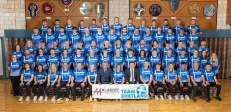 Group photo of Team Shetland in blue shirts, standing and seated in rows, holding a banner with sponsors "Malakoff Limited" and "Team Shetland." There are six plaques displayed on the wall behind them.