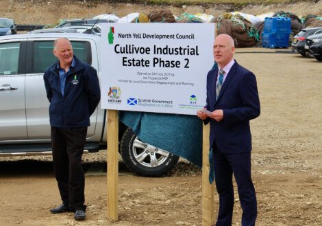 Two men stand beside a sign that reads "Cullivoe Industrial Estate Phase 2" at a development site. The sign notes a project by North Yell Development Council, Shetland Islands Council, HIE, and Scottish Government.