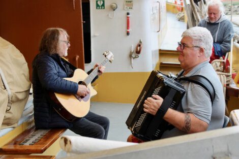 Two men playing musical instruments on a boat: one playing an acoustic guitar and the other playing an accordion. They are seated opposite each other near the railings.