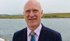 A bald man in a suit and tie stands outdoors near a body of water, with grassy terrain in the background.