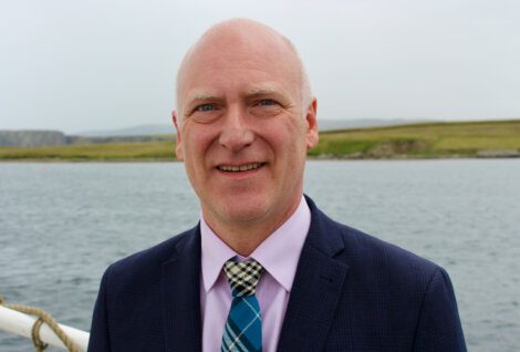 A bald man in a suit and tie stands outdoors near a body of water, with grassy terrain in the background.