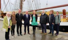 A group of eight people standing on a dock in front of a sailing ship on an overcast day. Seven are posing for the photo, and one appears to be walking slightly out of frame.