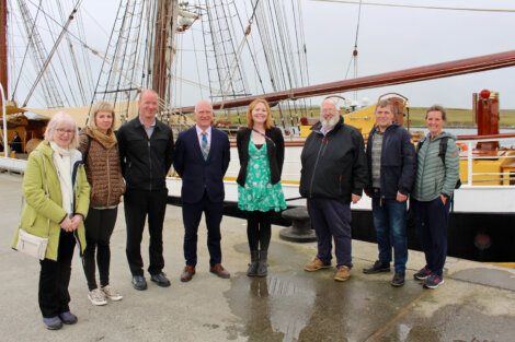 A group of eight people standing on a dock in front of a sailing ship on an overcast day. Seven are posing for the photo, and one appears to be walking slightly out of frame.