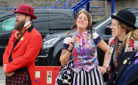 Three people dressed in colorful, quirky clothing stand outdoors, smiling and interacting with each other. There are cars and a blue staircase in the background.