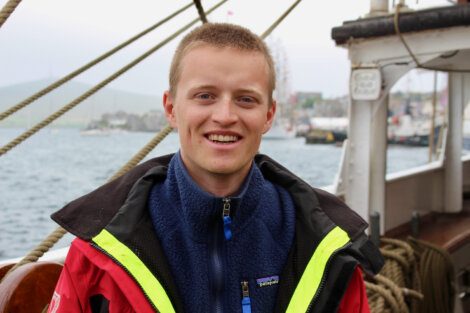 A person wearing a red and yellow jacket and a blue fleece stands on a boat. The background shows blurred water and harbor scenery.