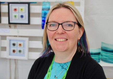 A person with glasses, wearing a black top and green lanyard, stands in front of artwork displayed on a white wall.