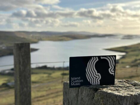 A sign reading "Island Custom Music" is propped on a wooden post with a scenic view of a lake and rolling hills in the background.